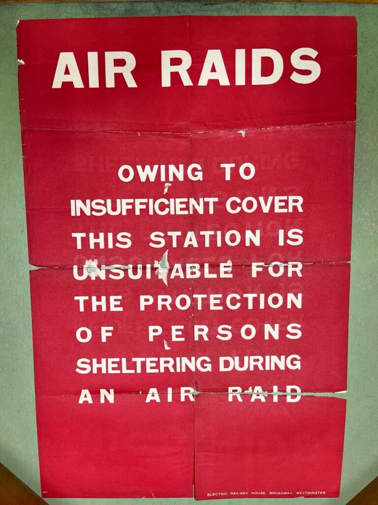 A printed sheet of paper with white text on a red background. The text reads: "AIR RAIDS
OWING TO
INSUFFICIENT COVER
THIS STATION IS
UNSUITABLE FOR
THE PROTECTION OF PERSONS SHELTERING DURING AN AIR RAID"