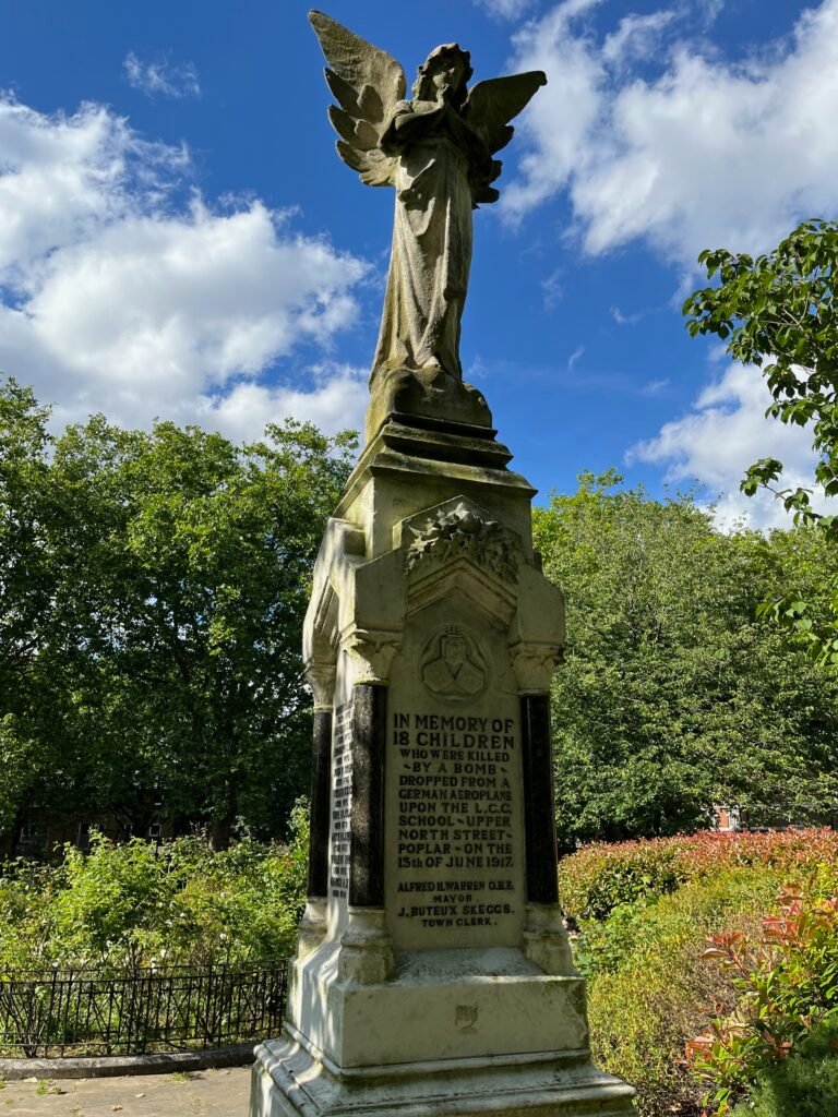 Memorial statue of an angel with inscriptions on the pedestal, set outdoors with greenery and a blue sky in the background.