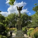 An angel statue with weathered stone texture stands in a sunlit garden, wings outstretched, amidst colourful flowers and greenery under a partly cloudy blue sky.