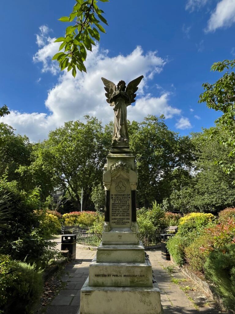 An angel statue with weathered stone texture stands in a sunlit garden, wings outstretched, amidst colourful flowers and greenery under a partly cloudy blue sky.