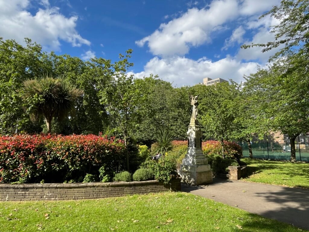 A serene park with a stone statue, red flowering shrubs, varied greenery, and a modern building in the distance under a partly cloudy sky.