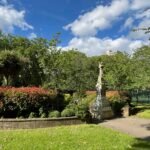 A serene park with a stone statue, red flowering shrubs, varied greenery, and a modern building in the distance under a partly cloudy sky.