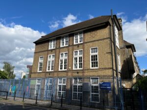 Three-story brick building with many white-framed windows and a steep roof, behind a tall blue metal fence, under a blue sky with clouds.