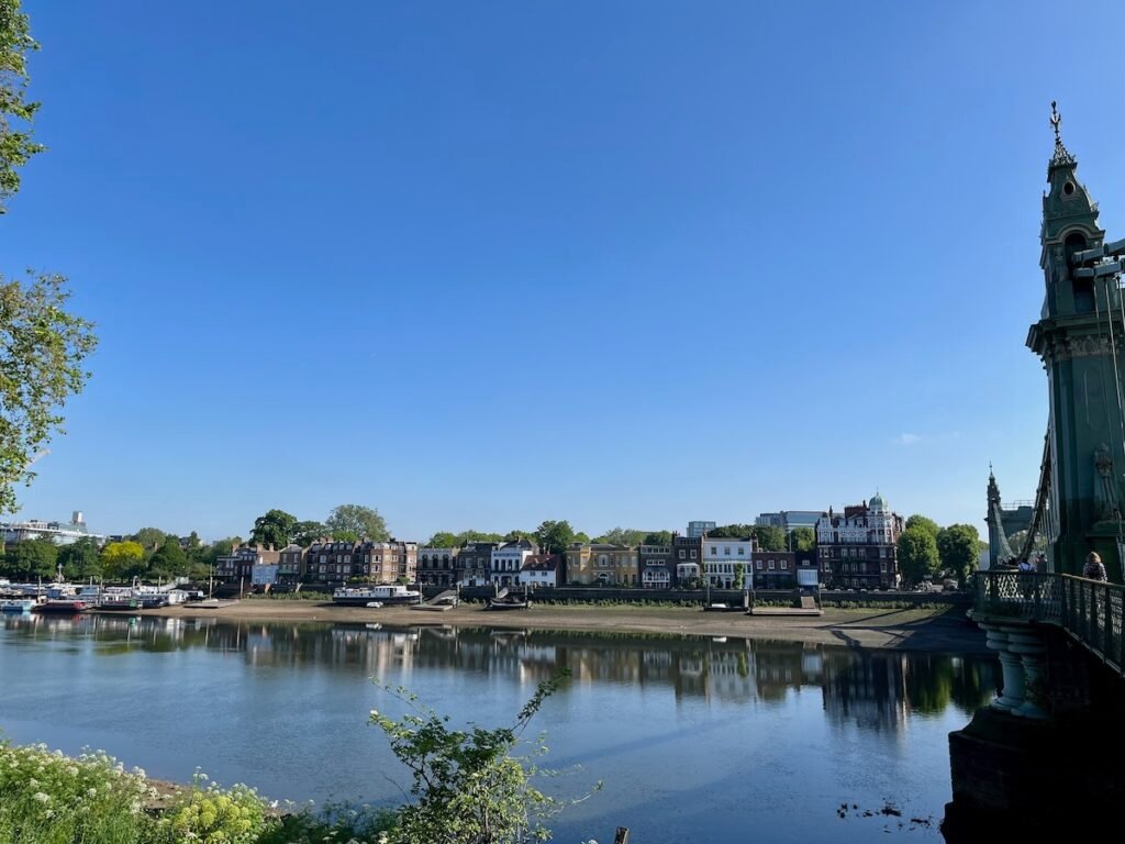 Scenic riverside view with buildings along the opposite bank, a few boats docked, and a section of an ornate bridge on the right under a clear blue sky.