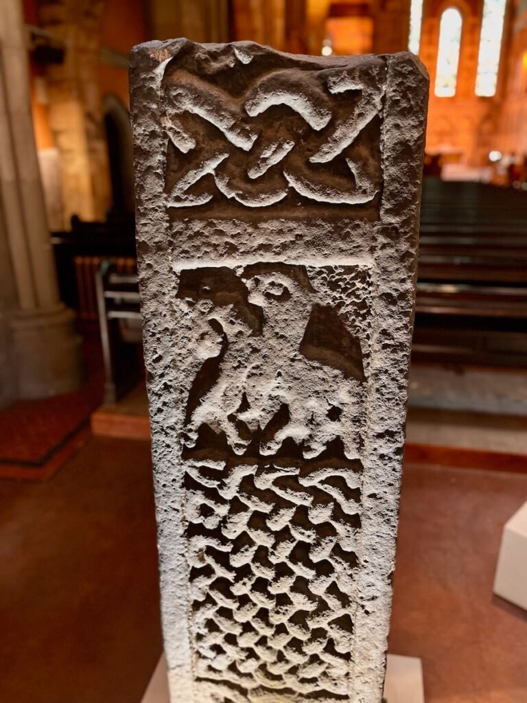 Carved stone pillar with knot design, abstract animal figure, and interwoven pattern, located inside a building with tall windows in the background.