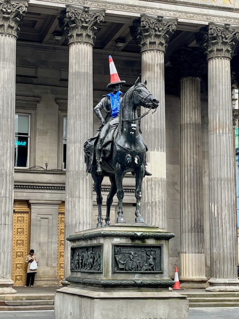 Statue of a man on a horse with a traffic cone on his head and a blue cloth around his shoulders in front of a neoclassical building.