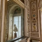 Ornately decorated room with a large mirror above a fireplace mantel reflecting a marble bust and curtains, surrounded by intricate gold and pastel wall details.