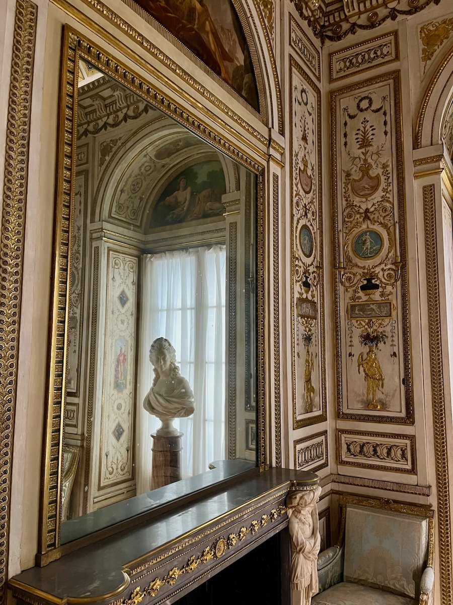 Ornately decorated room with a large mirror above a fireplace mantel reflecting a marble bust and curtains, surrounded by intricate gold and pastel wall details.