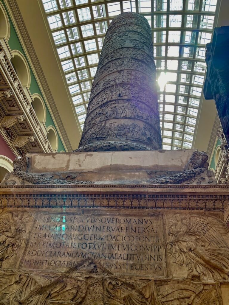 Detailed carved column with Latin inscriptions at its base, set in a historical building with an arched glass ceiling.