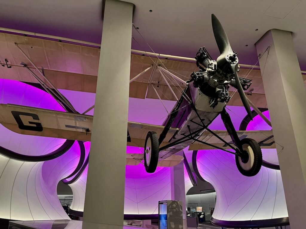 Vintage biplane suspended from the ceiling in a museum exhibit with magenta backlighting.