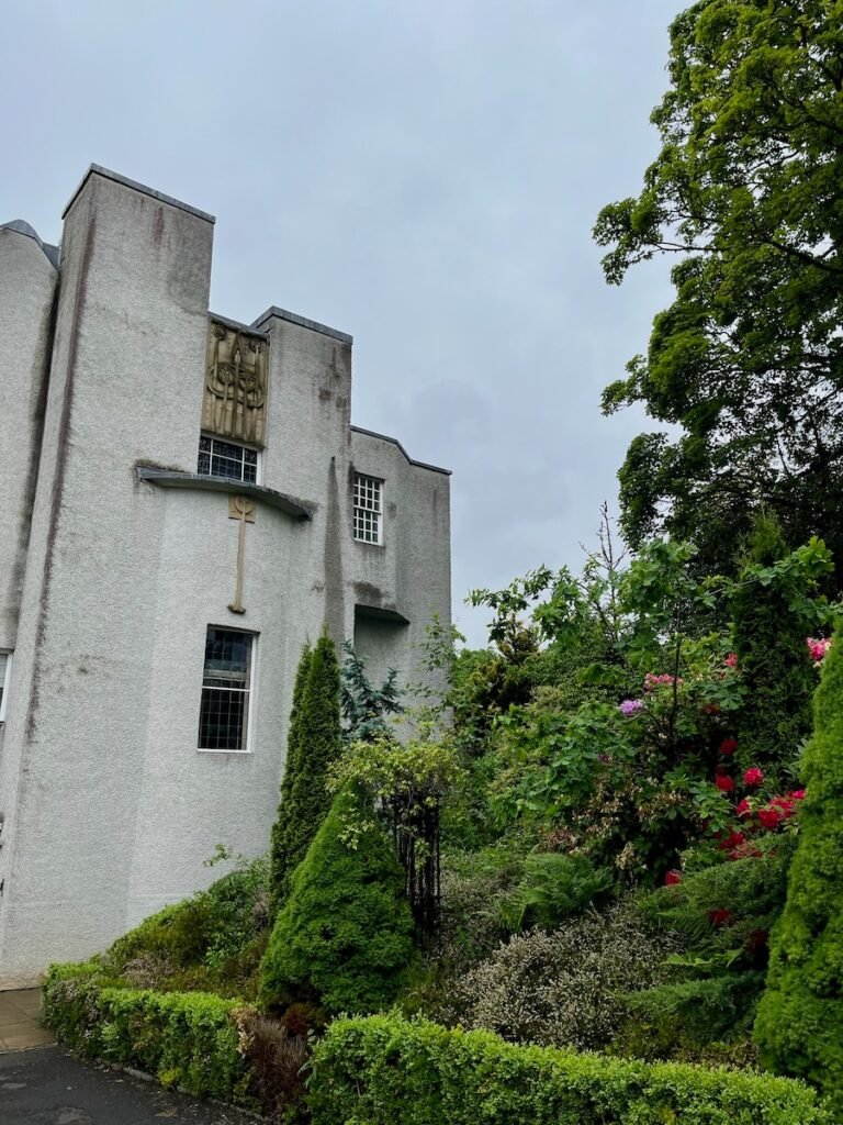 Grey stucco-finished building with a relief sculpture and lush greenery surrounding it.