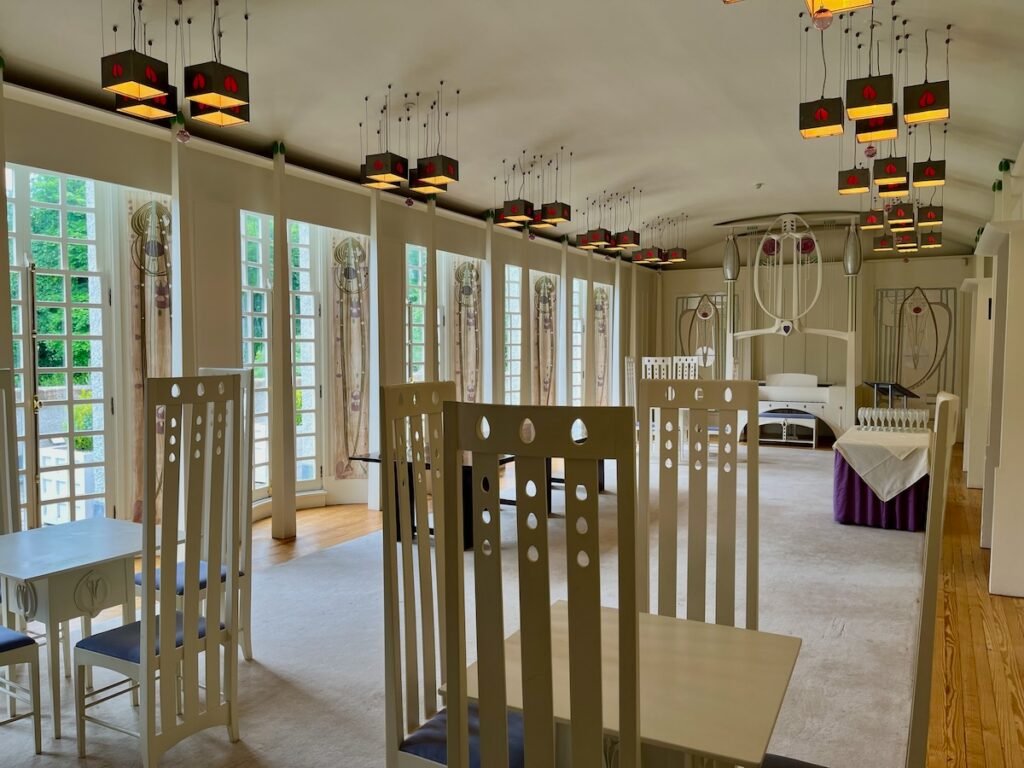 Art Nouveau-inspired room with tall white chairs, large windows, pendant lights, and abstract floral wall panels.