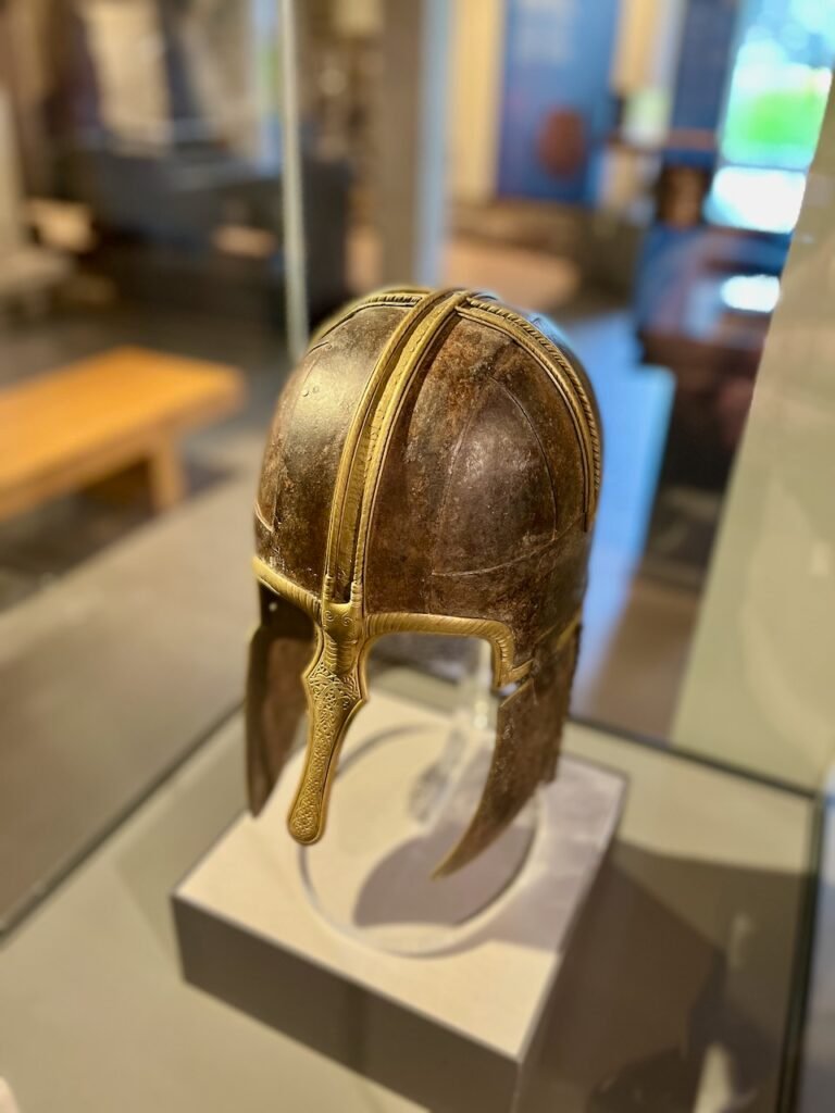 Ancient metal helmet with brass bands and engraved nose guard in a glass display case.