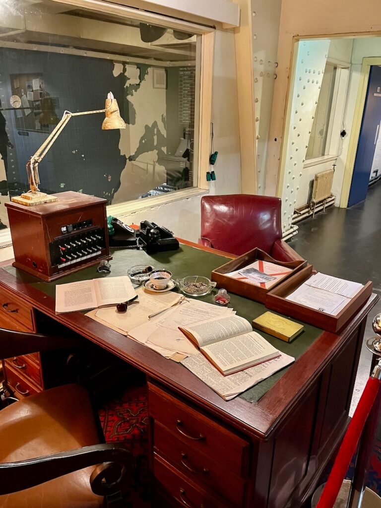 Vintage office desk with books, papers, a pipe, and a rotary phone, backed by a window showing a map.