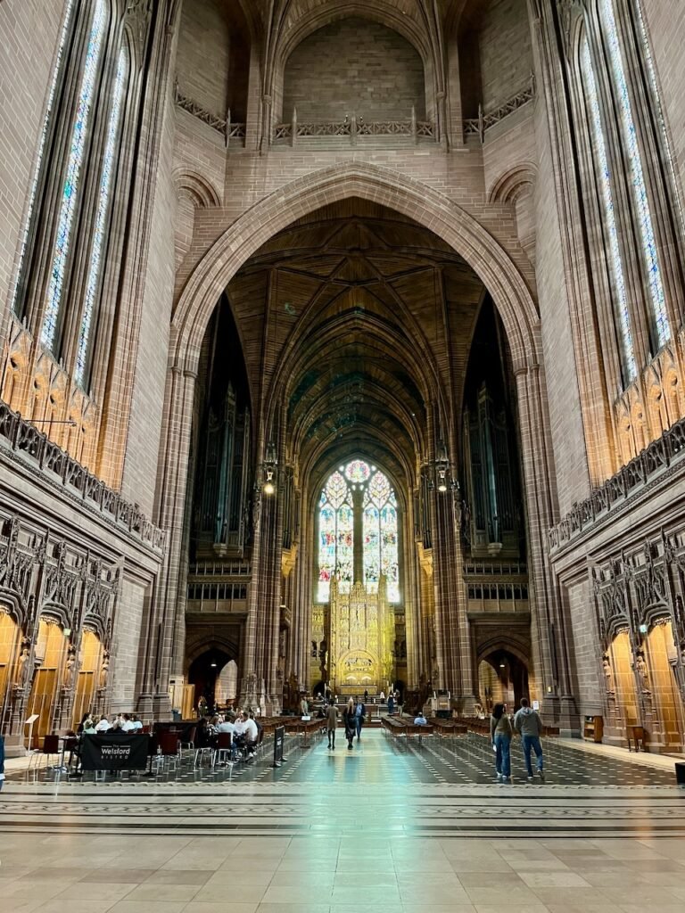 Interior of a large Gothic-style cathedral with a high arched entry, elaborate altar, and vibrant stained-glass windows. People are seated at a café on the left side.