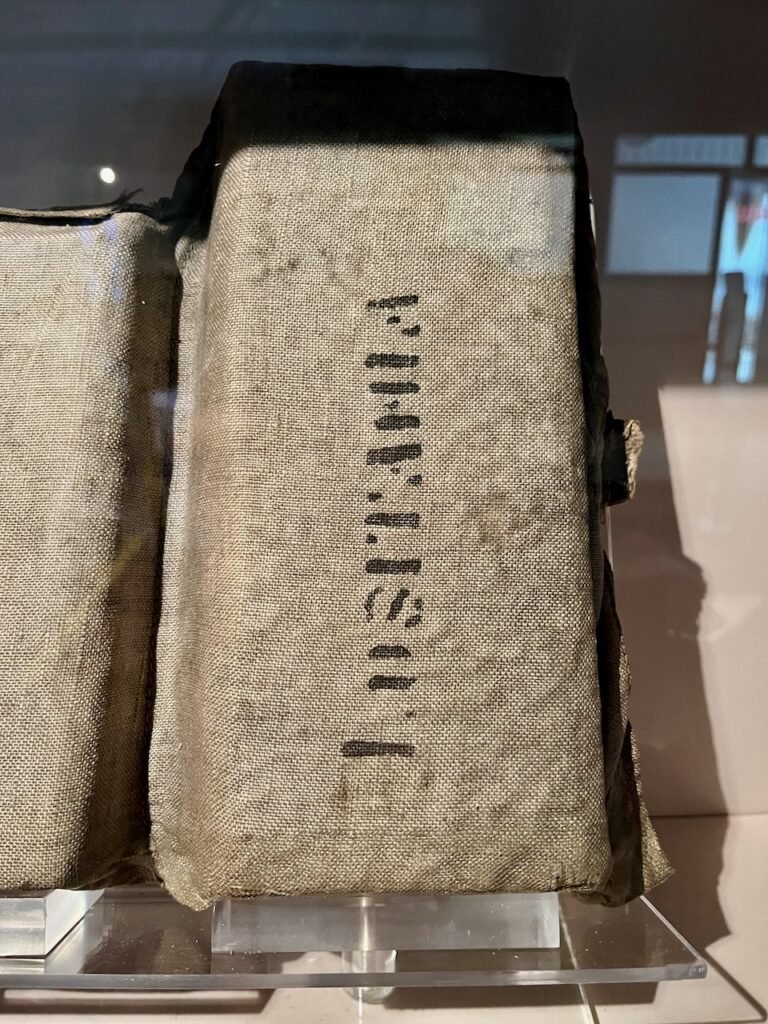 Close-up of a rectangular object wrapped in coarse, greyish-brown fabric with black printed text, supported by a transparent plastic stand.