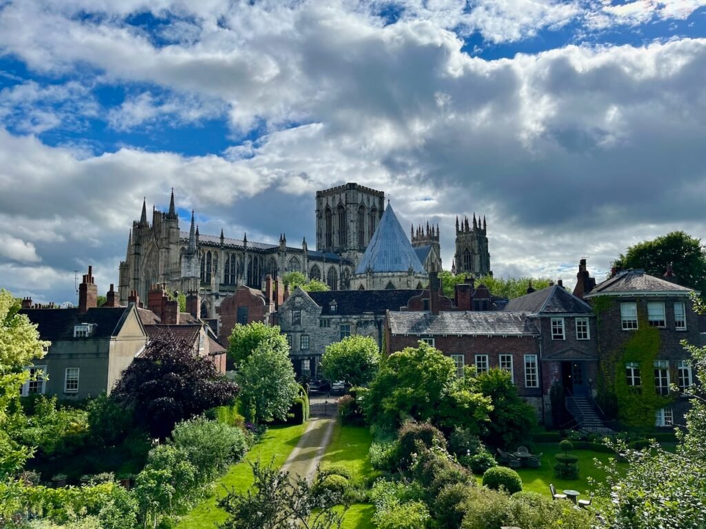 A Gothic cathedral behind traditional houses and gardens under a partly cloudy sky.