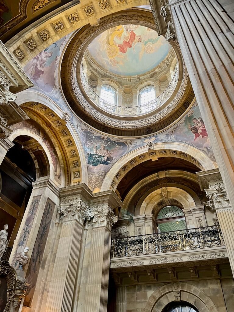 Ornate interior featuring a domed ceiling with classical frescos, arched windows, fluted Corinthian columns, and an intricate wrought-iron balcony.