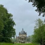 A small stone building with a domed roof and portico, surrounded by dense trees and accessed via a grassy path under an overcast sky.