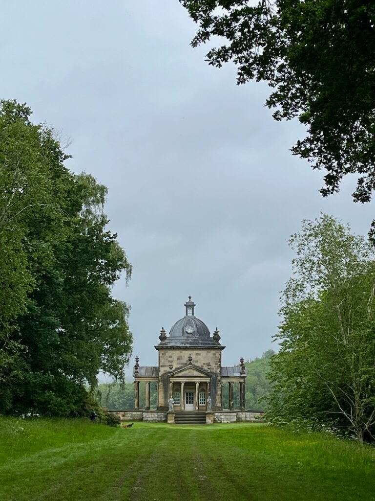 A small stone building with a domed roof and portico, surrounded by dense trees and accessed via a grassy path under an overcast sky.