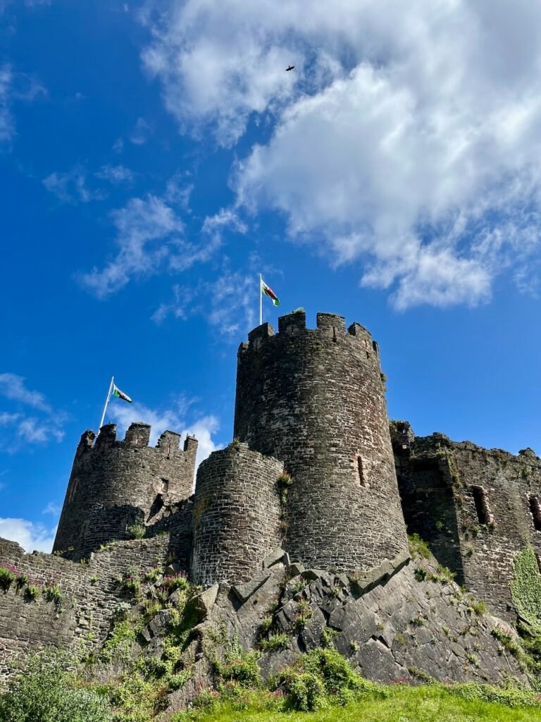 A historic stone castle with two round towers and flags, set against a blue sky with clouds.