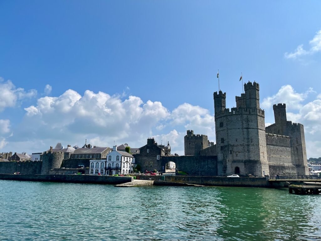 Stone castle by water with towers and adjacent historical buildings against a bright blue sky.