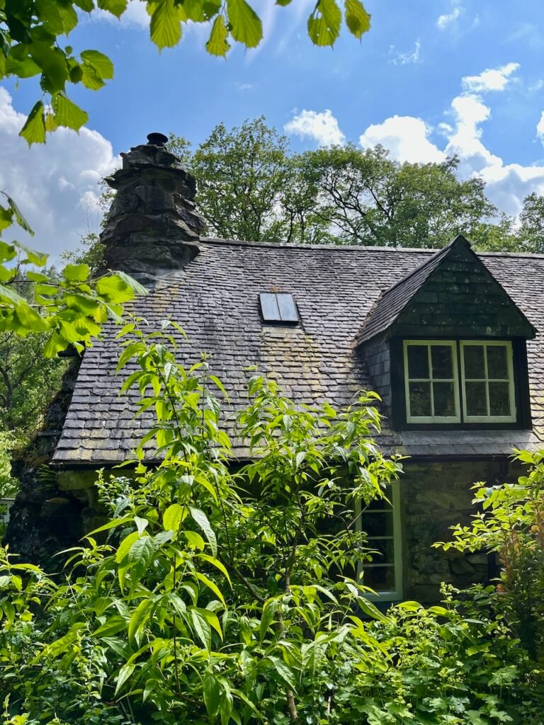 A stone cottage with a slate roof and chimney, surrounded by lush greenery under a blue sky with clouds.