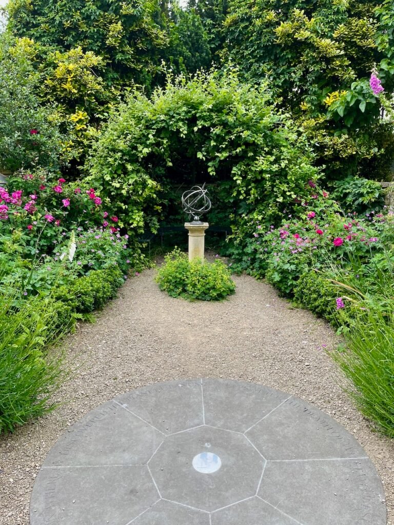 Garden with armillary sphere on a pedestal, surrounded by lush greenery and pink flowers.