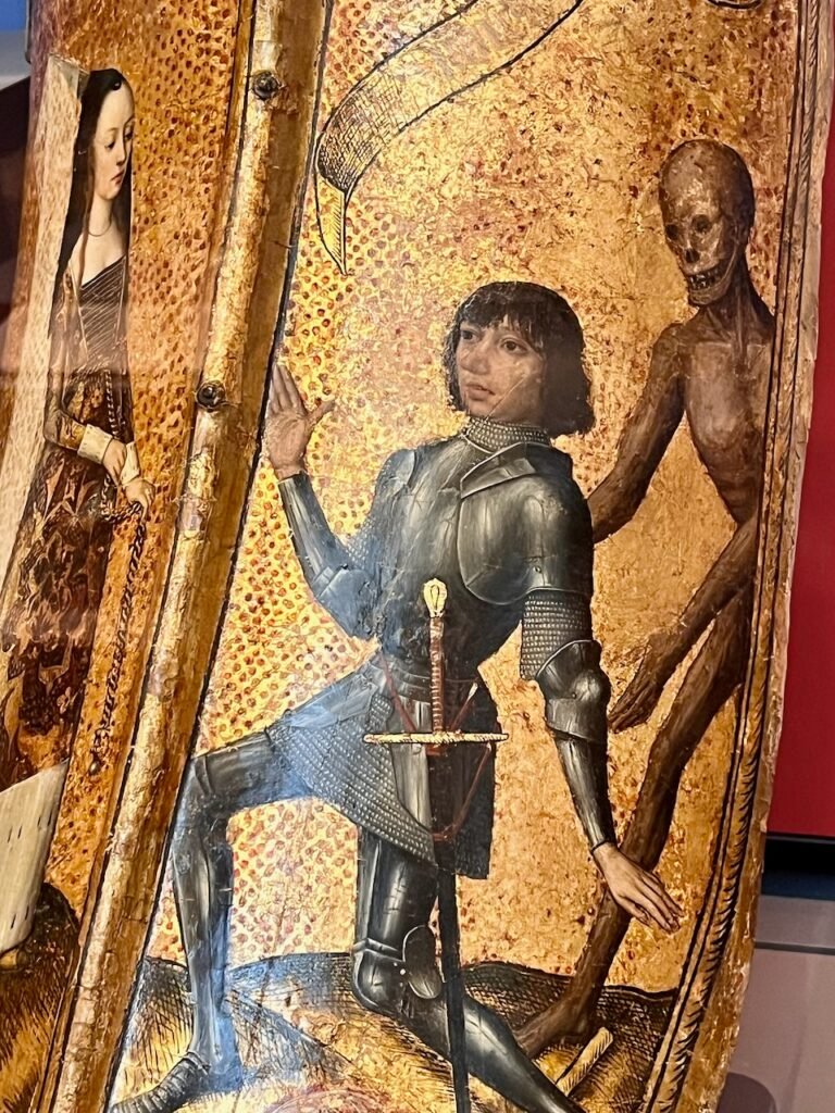 Scene from a shield showing a knight kneeling before a lady, with Death standing behind him.