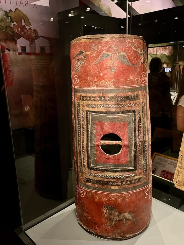 A large ancient cylindrical red shield with painted winged figures, geometric motifs, and a lion, displayed in a museum.