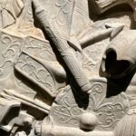 Close-up of a stone relief sculpture depicting intricately carved armour and shields with floral patterns.