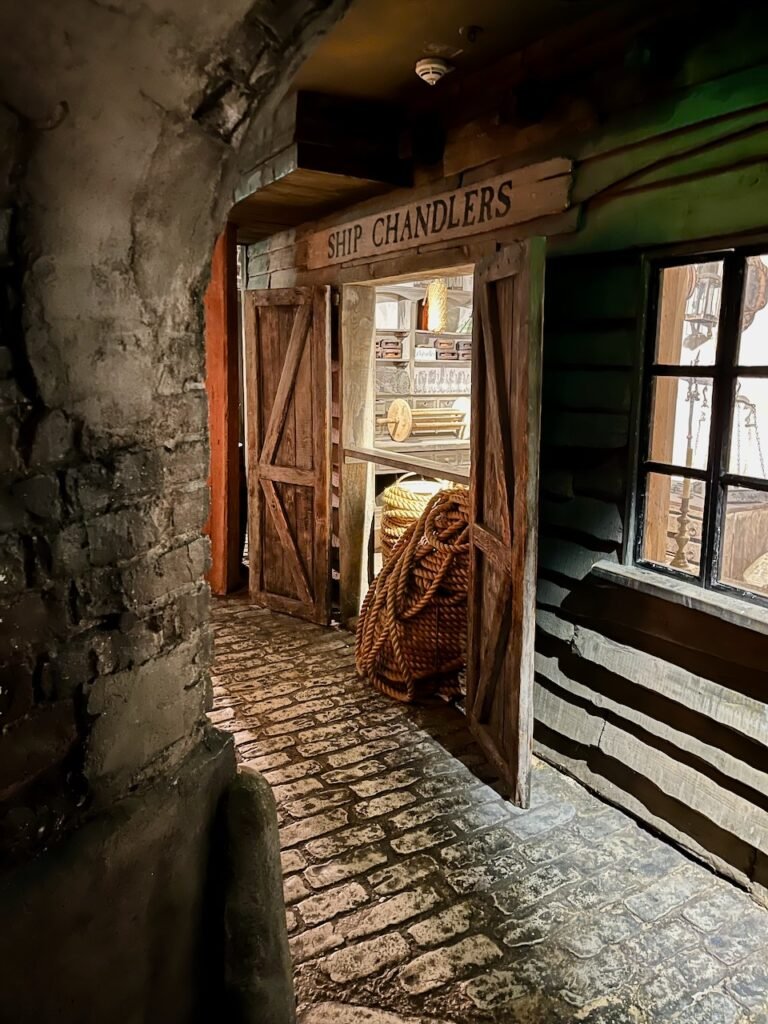 Entrance to a rustic ship chandler's shop with wooden doors and cobblestone pathway.