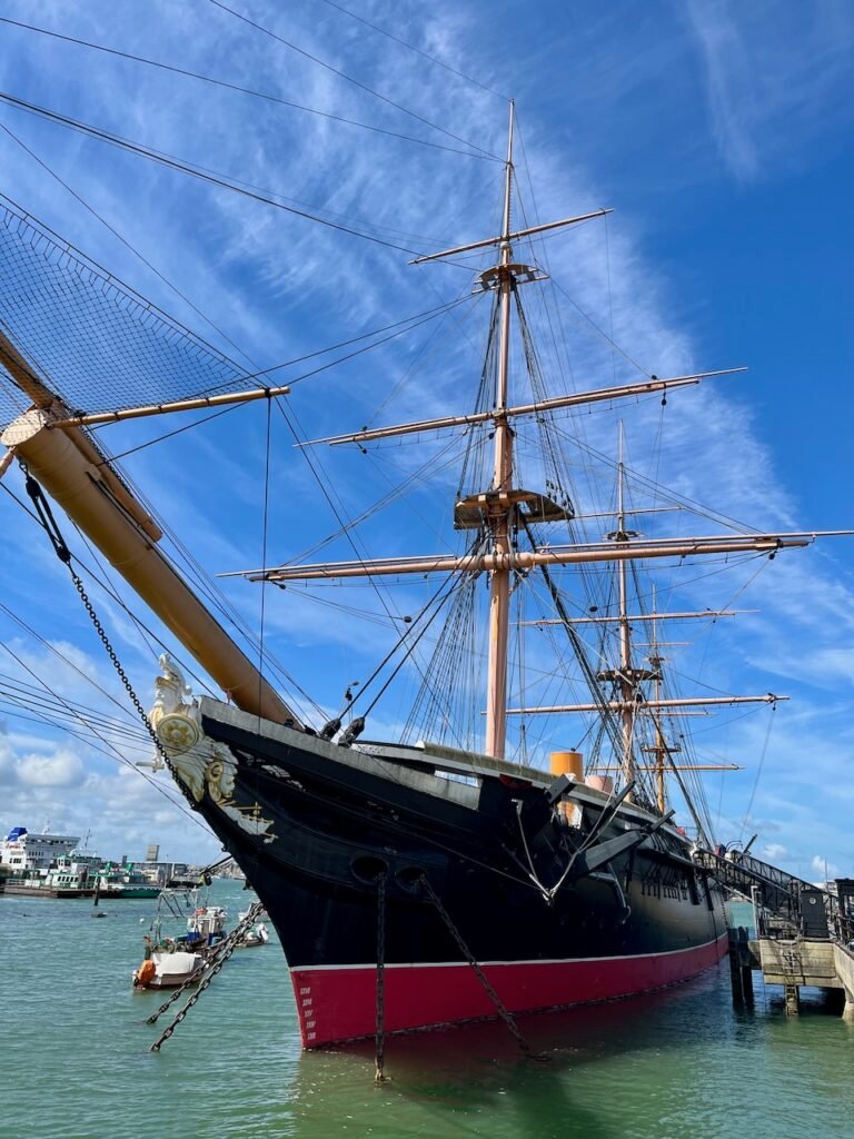 Historic ship with black hull and red stripe docked at a pier under a blue sky.