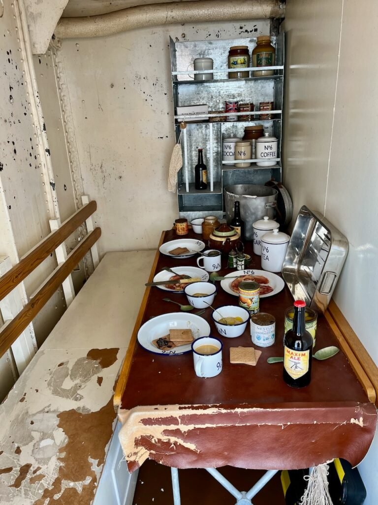 A worn pantry with a table set with enamelware and food items, surrounded by shelves filled with jars and bottles.