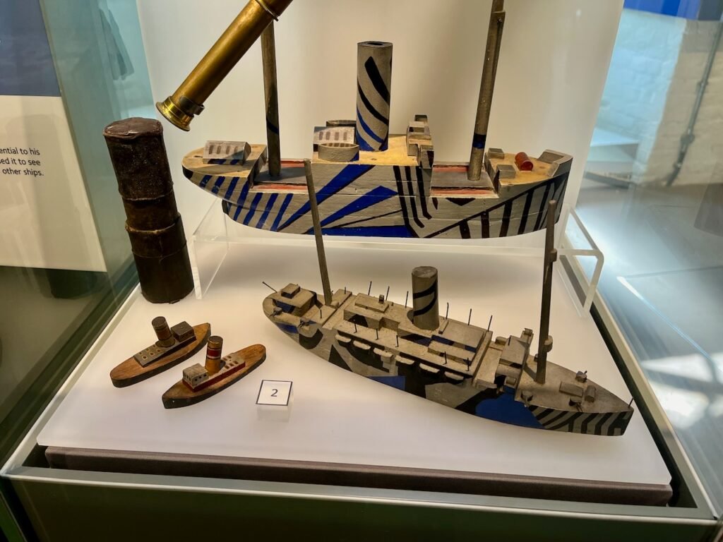 Display case with dazzle-patterned ship models and a telescope.