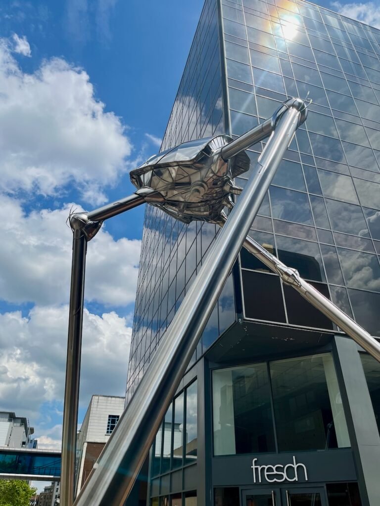 Metallic sculpture in front of a glass building with a blue sky and clouds.