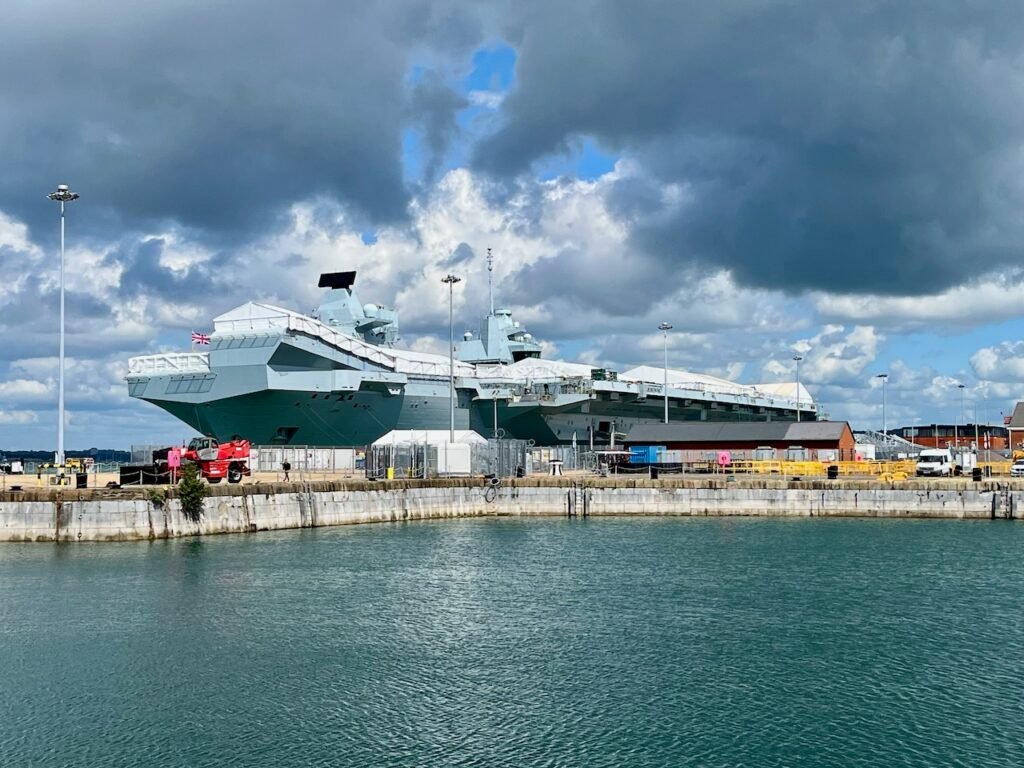 A big aircraft carrier, docked, with a green expanse of water in the foreground and dark clouds above.