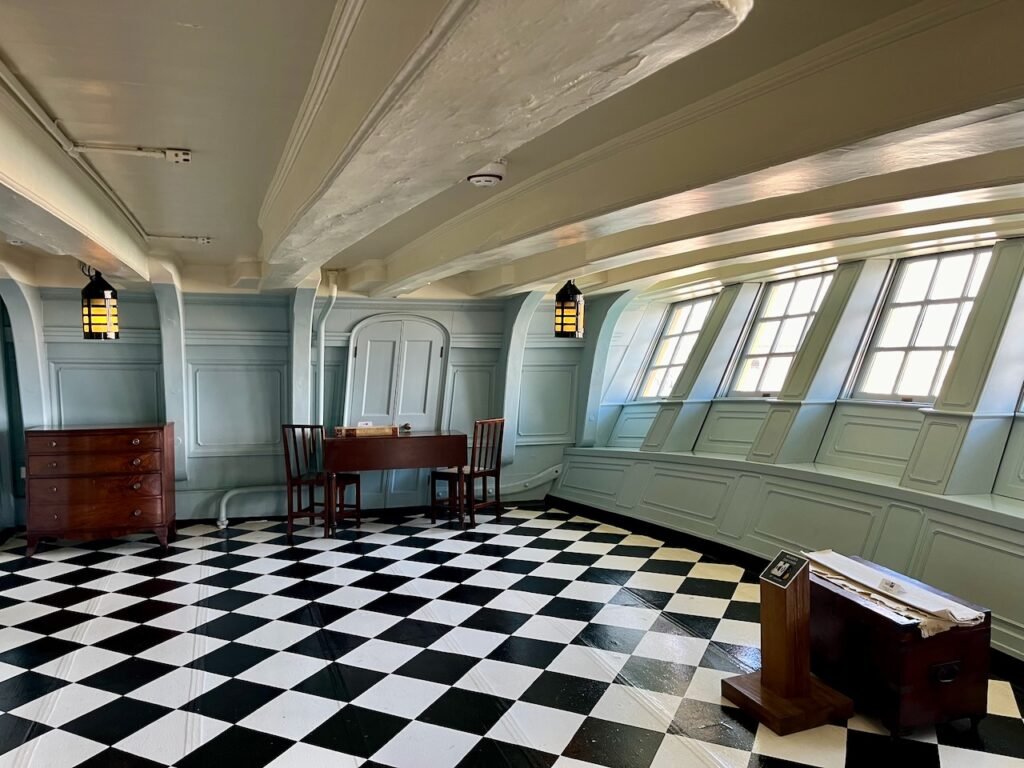 A big room in a wooden ship, with external windows on the slanted wall to the right and a black and white chequered floor pattern.