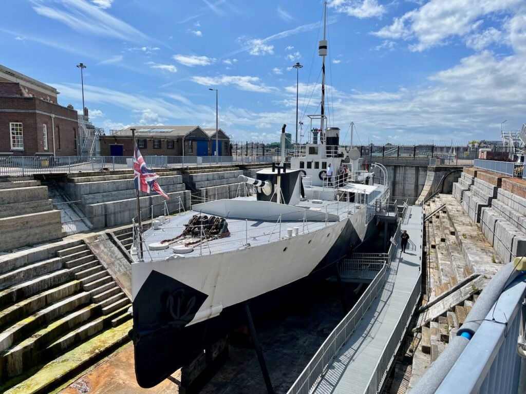 A decommissioned warship with a Union Jack in a dry dock surrounded by historical brick buildings under a blue sky.