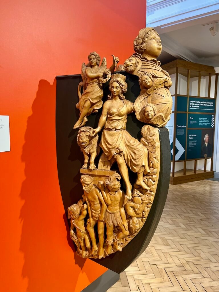 A detailed wooden sculpture with classical figures against a red wall in a museum.