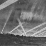 Aurorae at war The image depicts a night scene where several beams of light, likely from searchlights, intersect against a cloudy sky. The beams create a crisscross pattern, illuminating portions of the dark, overcast sky. A few stars are visible amidst the clouds, adding to the atmospheric effect. The bottom of the image shows a silhouetted horizon, possibly of a landscape with scattered trees and buildings. The overall tone is monochromatic, with shades of grey highlighting the dramatic effect of the light beams against the night sky.