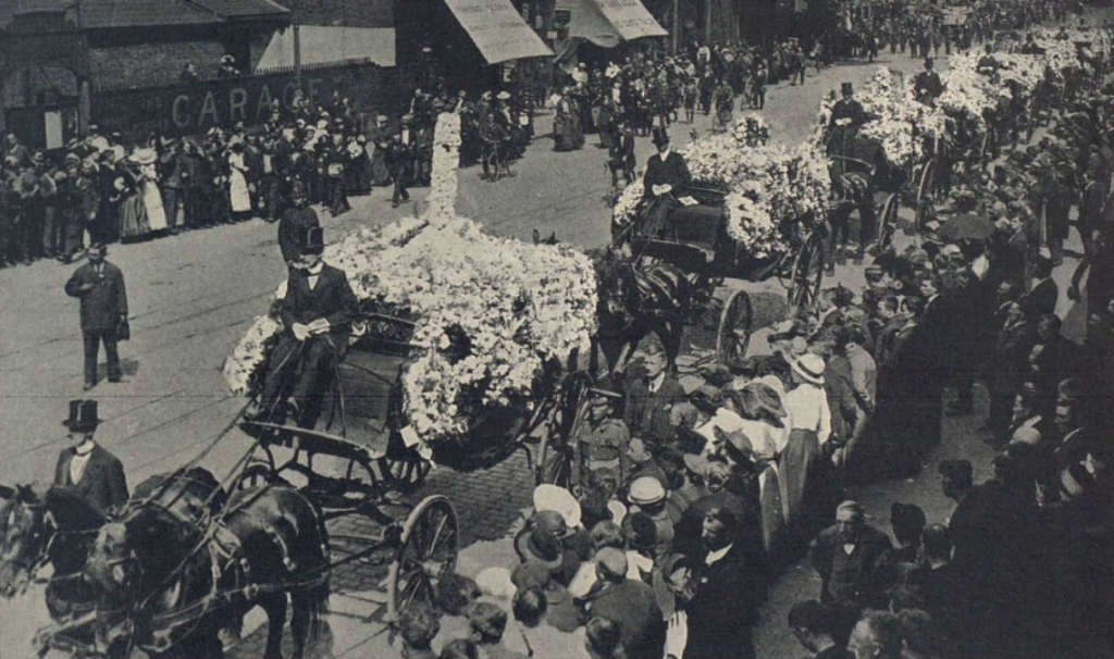 A historical funeral procession with horse-drawn carriages decorated with flowers, moving along a cobblestone street watched by a crowd.