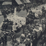 A historical funeral procession with horse-drawn carriages decorated with flowers, moving along a cobblestone street watched by a crowd.