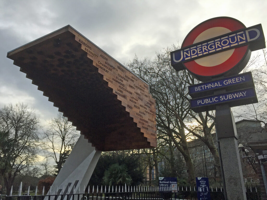 Stairway to Heaven Memorial, Bethnal Green, with engraved names on a cantilevered roof, supported by a white concrete base, surrounded by bare trees. To the right, a traditional London Underground sign displays "UNDERGROUND," "BETHNAL GREEN," and "PUBLIC SUBWAY."