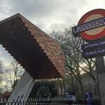 Stairway to Heaven Memorial, Bethnal Green, with engraved names on a cantilevered roof, supported by a white concrete base, surrounded by bare trees. To the right, a traditional London Underground sign displays "UNDERGROUND," "BETHNAL GREEN," and "PUBLIC SUBWAY."