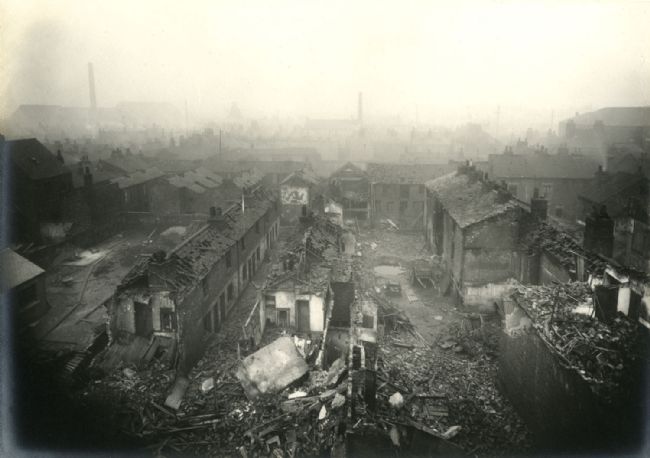 A black-and-white photograph shows a devastated urban neighbourhood viewed from an elevated position. Rows of densely packed buildings line a central street, with many roofs collapsed and walls partially destroyed. Rubble, broken timber and masonry fill the foreground and extend along the street. Smoke or haze hangs over the scene, obscuring a distant industrial skyline with chimneys and tall structures. 